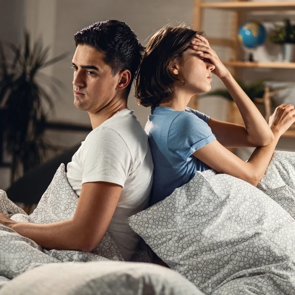 Man and woman sitting on a bed with their backs together with a worried look on their faces.